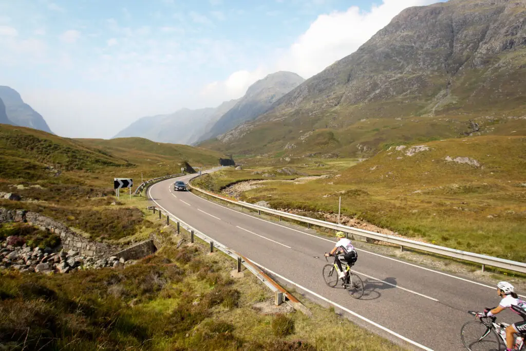 Cyclists on a scenic road through the rugged landscapes of the Scottish Highlands, showcasing beautiful mountain views perfect for exploring on a Scotland self-drive tour.