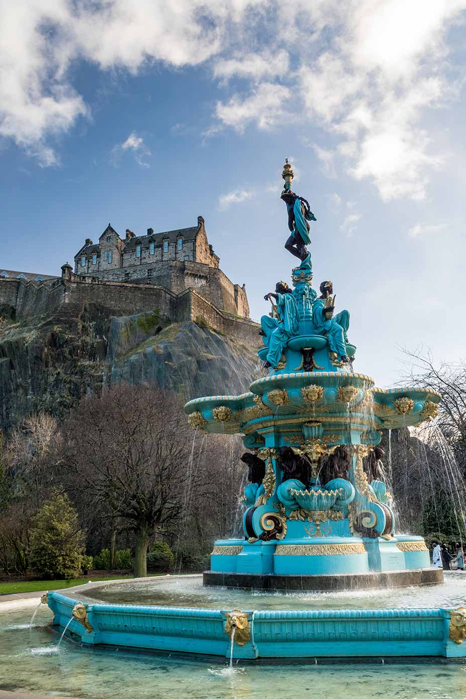 Ross Fountain with vibrant turquoise and gold colors in the foreground, and the historic Edinburgh Castle sitting atop a rocky hill in the background, captured in West Princes Street Gardens, Scotland.