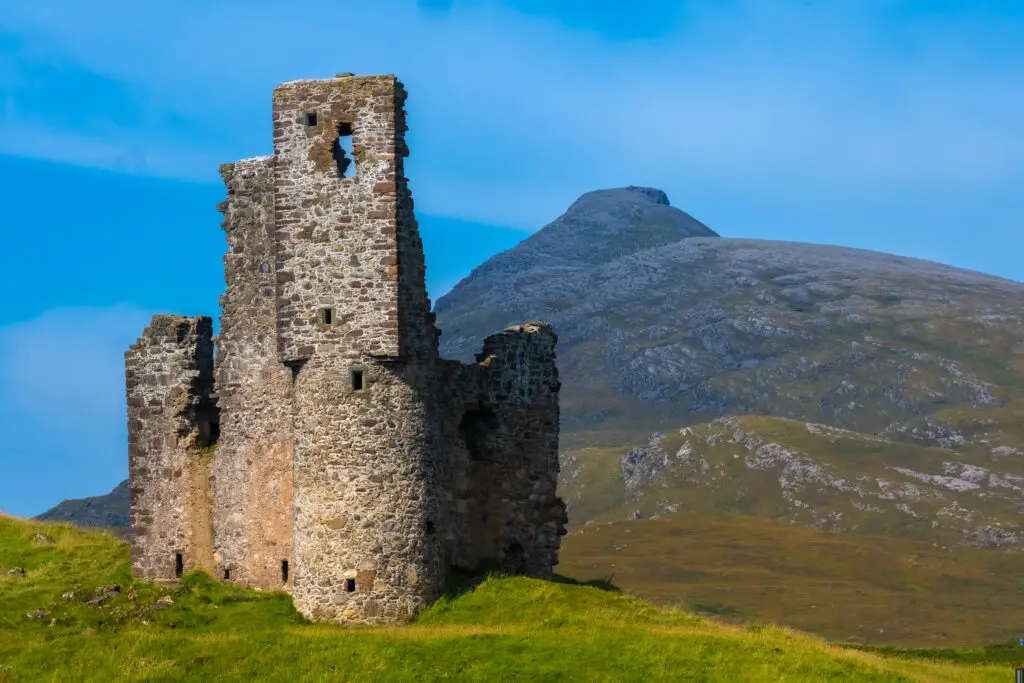 Ardvreck Castle ruins on a rocky promontory at Loch Assynt, surrounded by lush grass and mountain backdrop, in Sutherland, Scottish Highlands.